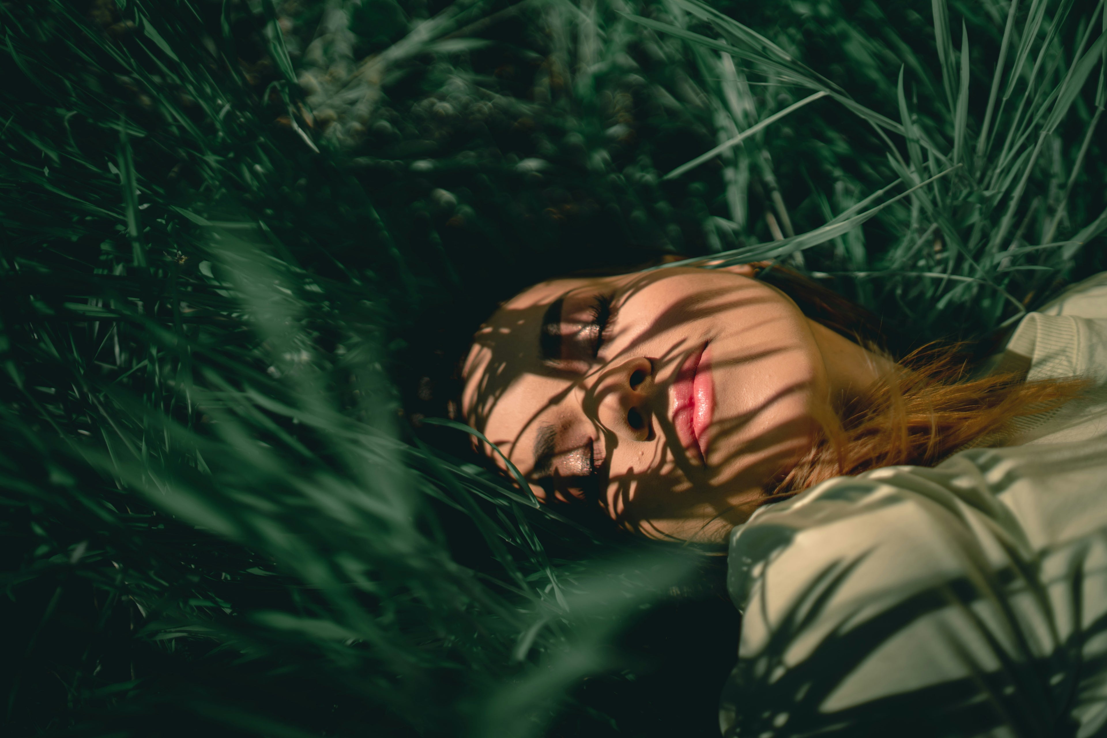 Woman lying in tall grass with sunlit lips, wearing Ocean Avenue Beauty Peel-Off Lip Stain—natural outdoor lifestyle shot.